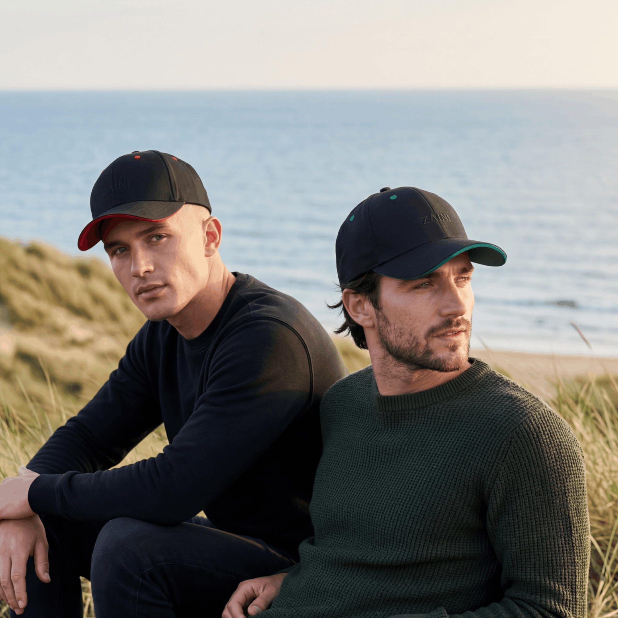 Two men sitting on a beach wearing baseball caps with ocean in the background