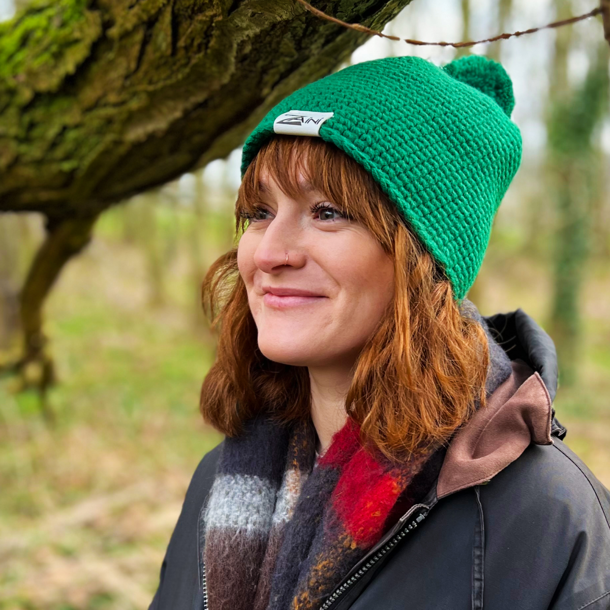 A woman wearing a green fleece lined snug bobble hat with the brand logo visible on the hat's fold.