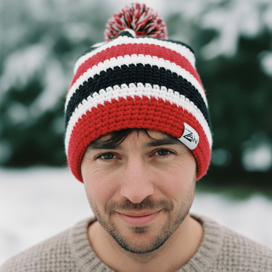 Man wearing a red, white, and black striped knit beanie with a logo in a snowy outdoor setting.