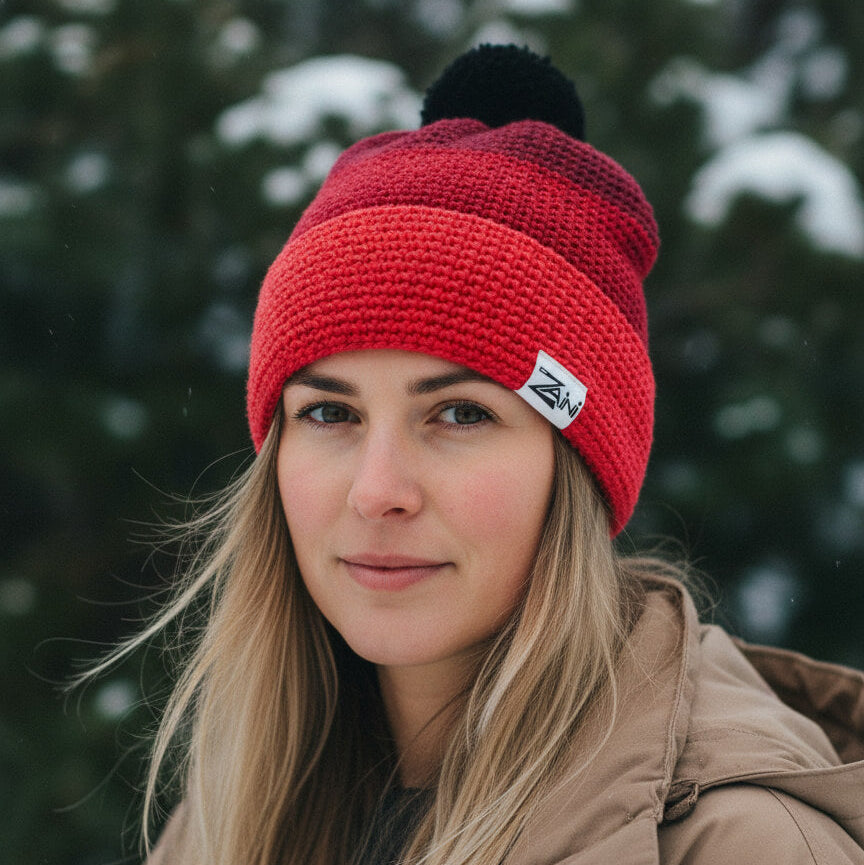Woman wearing a red knit beanie with a black pom-pom and a logo, standing in a snowy forest.