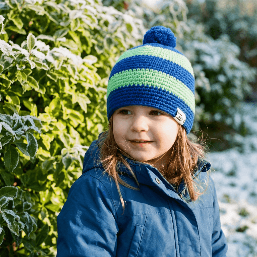 Child wearing a blue and green striped knit hat with a pom-pom, standing in a snowy landscape.