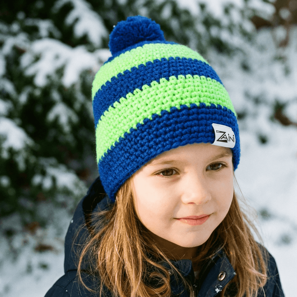 Child wearing a blue and green striped knit hat with a brand logo, standing in a snowy outdoor setting.
