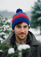 Man wearing a blue and red beanie with a pom-pom, standing outdoors with snow-covered trees in the background.