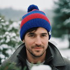 Man wearing a blue and red beanie with a pom-pom, standing outdoors with trees in the background.
