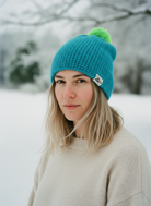 Person wearing a blue knit beanie with a green pom-pom in a snowy landscape