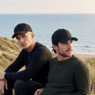Two men sitting on a beach wearing baseball caps with ocean in the background