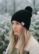 Person wearing a black knit beanie with a brand logo in a snowy forest setting