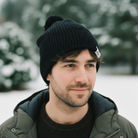 Man wearing a black knit beanie and green jacket in a snowy landscape