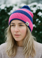 Person wearing a pink and blue striped knit beanie with a brand logo, standing outdoors with blurred greenery in the background.