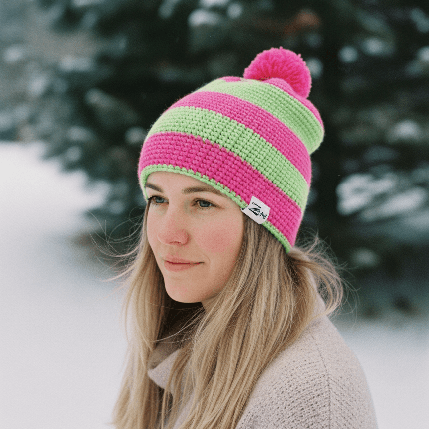 Person wearing a pink and green striped beanie with a pom-pom in a snowy outdoor setting.