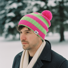Man wearing a pink and green striped beanie with a pom-pom in a snowy landscape.