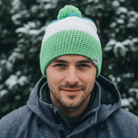 Man wearing a green and white beanie with a pom-pom in front of snow-covered trees.