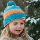 Child wearing a blue and orange striped knit hat with a pom-pom, standing in a snowy forest.