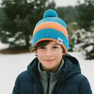 Child wearing a blue and orange striped knit hat in a snowy landscape with trees.