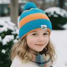 Child wearing a blue and orange knit hat with a brand logo, standing in a snowy outdoor setting.