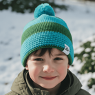 Child wearing a green and blue knit beanie with a pom-pom in a snowy outdoor setting