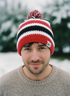 Man wearing a red, white, and black striped knit beanie with a brand logo outdoors.