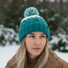 Person wearing a teal knit beanie with a pom-pom in a snowy outdoor setting