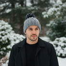 Man wearing a gray knit beanie and black coat standing in a snowy landscape with trees.