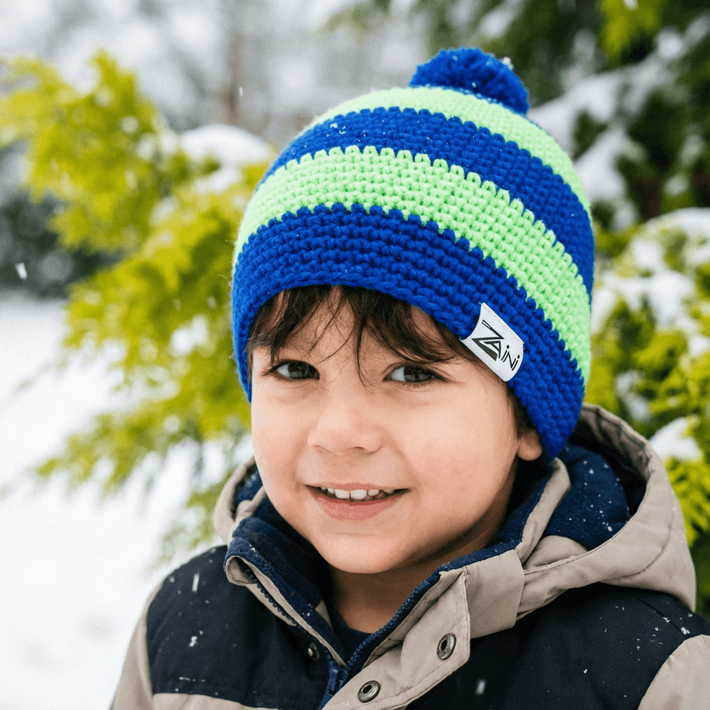 Child wearing a blue and green striped knit hat with a brand logo, standing outdoors in a snowy landscape.